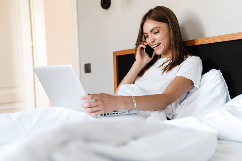 Mujer joven atractiva usando el teléfono móvil y el ordenador portátil mientras se relaja en la cama - Foto, imagen