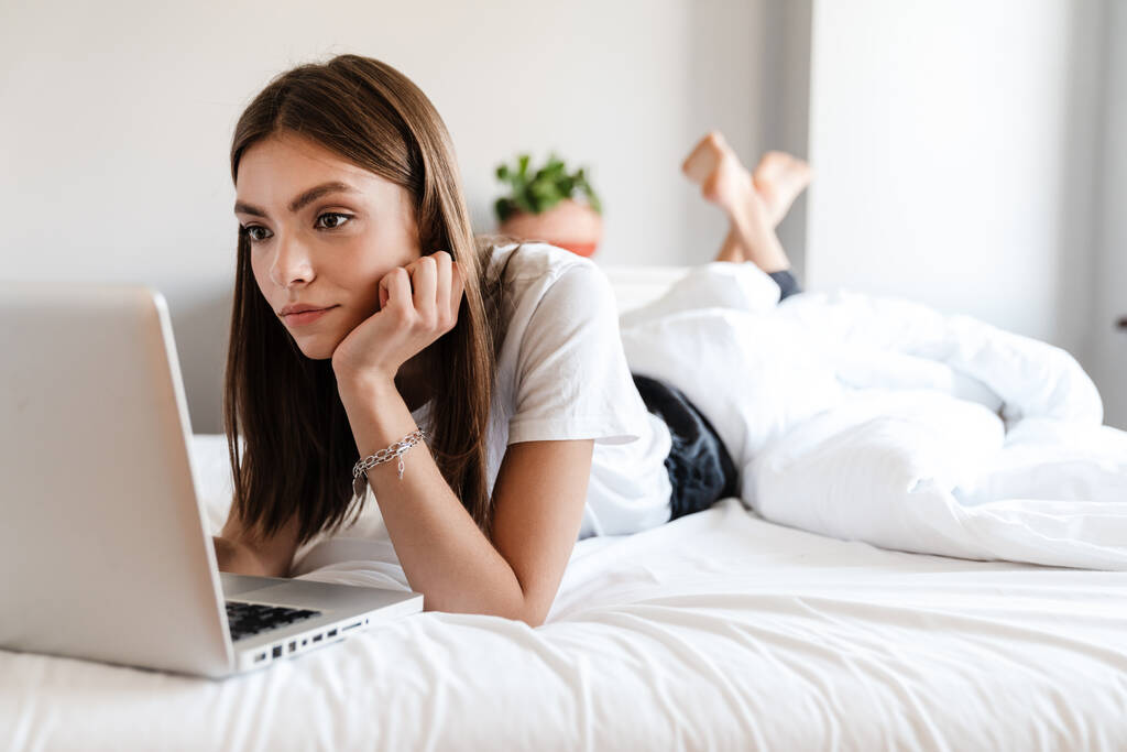 Ελκυστική νεαρή γυναίκα που χρησιμοποιεί φορητό υπολογιστή ενώ χαλαρώνει στο κρεβάτι - Φωτογραφία, εικόνα