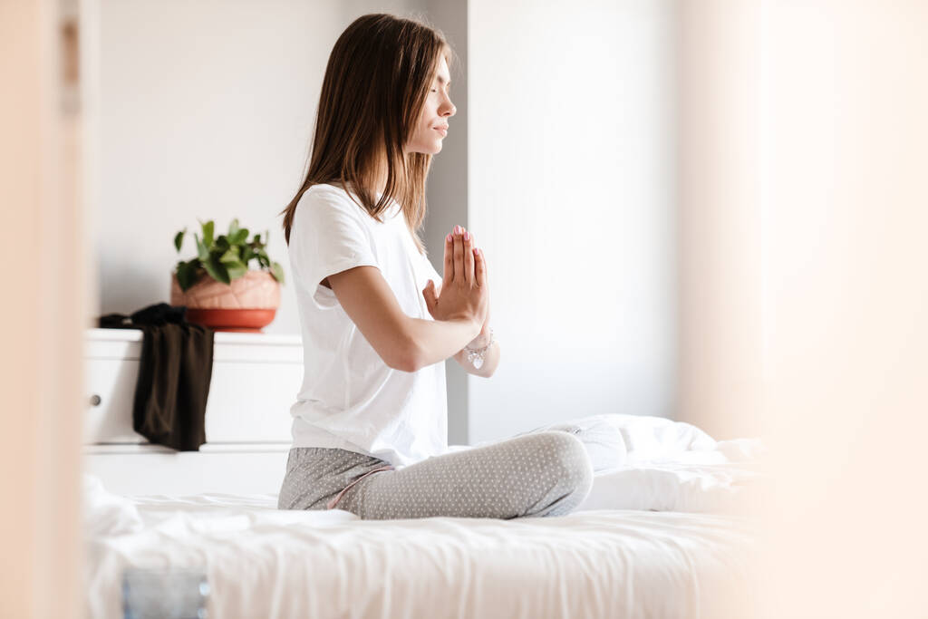 Молодая женщина медитирует с закрытыми глазами в яркой спальне, сидя на кровати - Фото, изображение