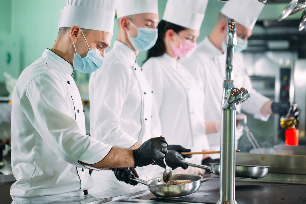 Chefs in protective masks and gloves prepare food in the kitchen of a restaurant or hotel. - Photo, Image