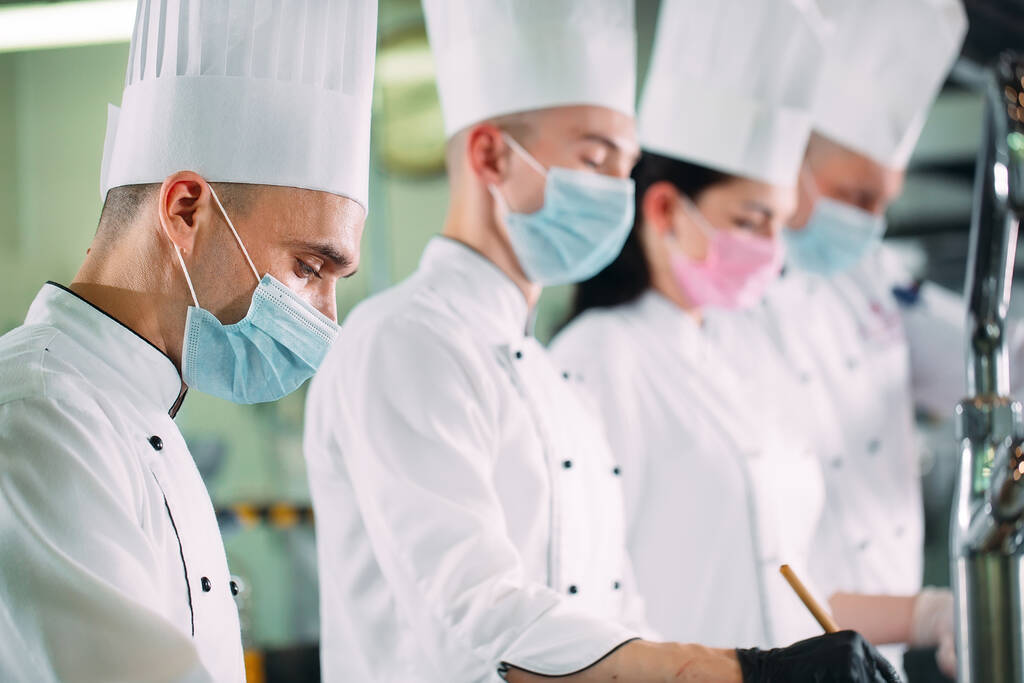 Chefs con máscaras protectoras y guantes preparan comida en la cocina de un restaurante u hotel. - Foto, imagen