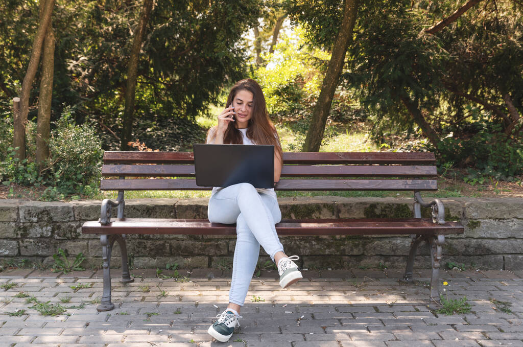 Young woman sitting in the park with laptop talking with operator over smartphone and typing instruction - Photo, Image