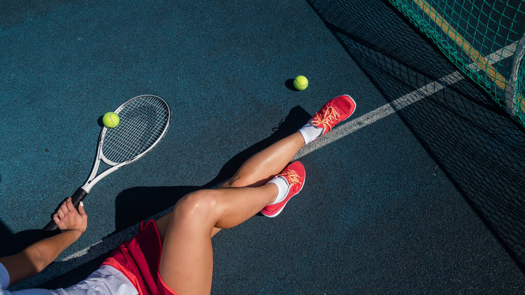 Spor etekli yüzü olmayan bir kız tenis kortunda oturuyor ve elinde bir roket tutuyor. Kadın bacaklarının üst görüntüsü. - Fotoğraf, Görsel