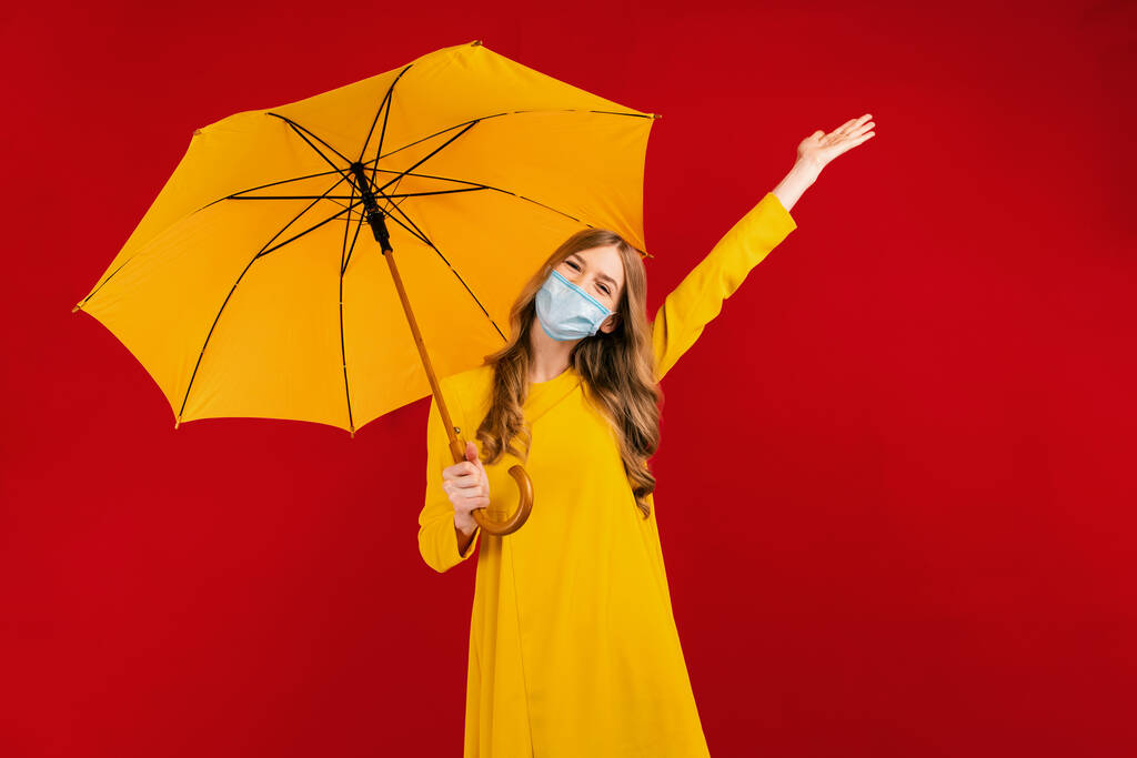 Ευτυχισμένο κορίτσι με ένα κίτρινο φόρεμα και μια ιατρική προστατευτική μάσκα στο πρόσωπό της διασκεδάζει, με μια ομπρέλα στα χέρια της - Φωτογραφία, εικόνα