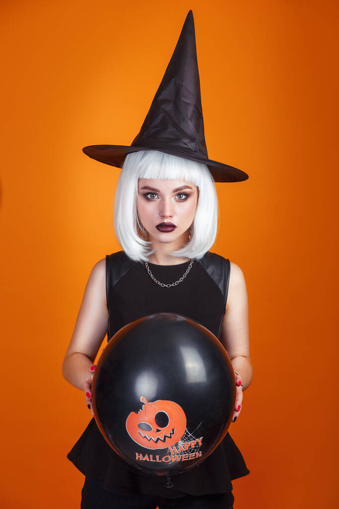 Fiesta de Halloween. Feliz Halloween sexy bruja con pelo blanco sosteniendo negro y naranja Globo de aire con calabaza sobre fondo naranja. - Foto, Imagen