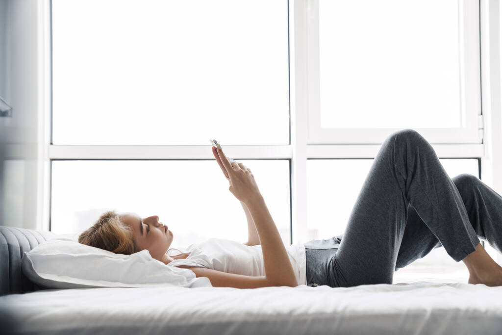 Foto de una joven enfocada usando un teléfono celular mientras está acostada en la cama después de dormir en una habitación luminosa - Foto, imagen