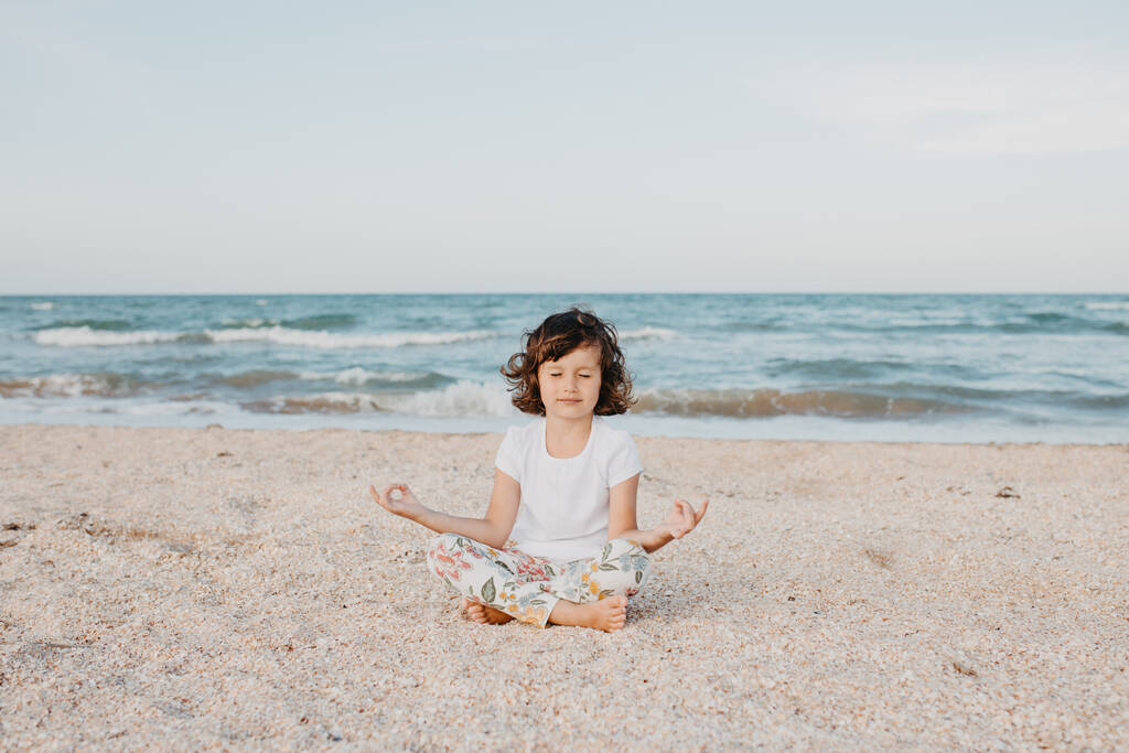 Dziecko dziewczyna siedzi na plaży medytując, uprawiając jogę, zrelaksować uważny oddech - Zdjęcie, obraz