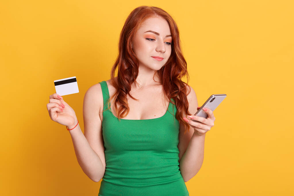 Imagem de mulher linda com cabelo ruivo vestindo roupas casuais, segurando cartão de crédito e celular, olhando para a tela do dispositivo, fazendo compras on-line. - Foto, Imagem
