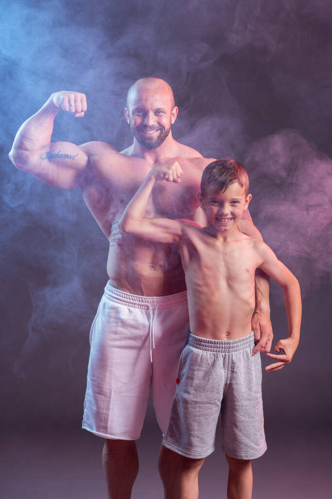 Potężny muskularny mężczyzna bez koszulki pozujący z synem w studio z czerwonym i niebieskim dymem - Zdjęcie, obraz