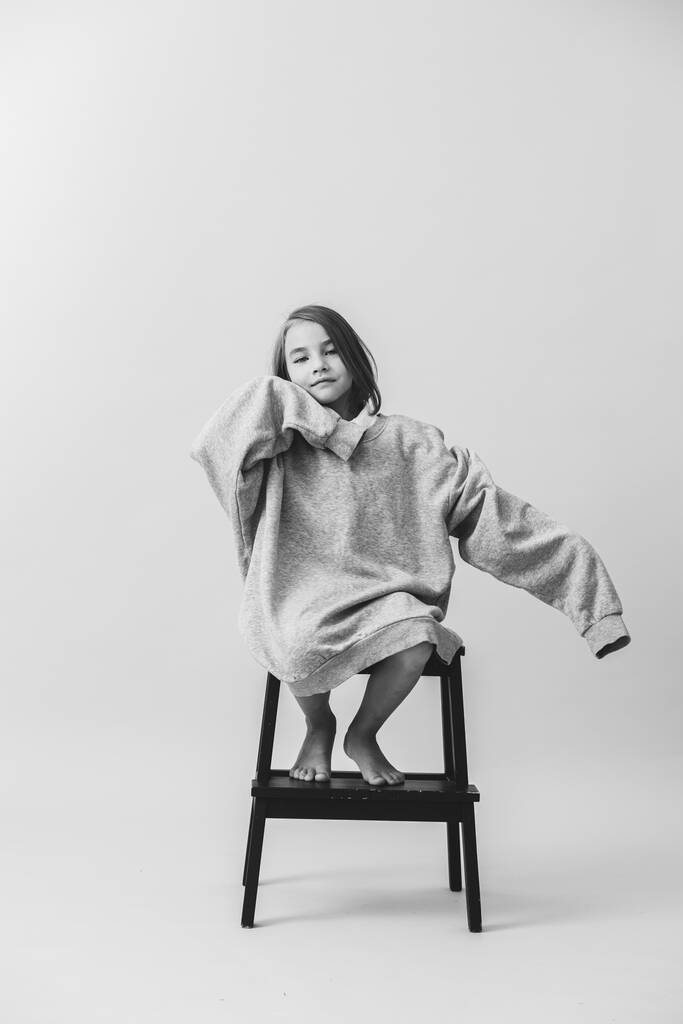 Czarno-białe zdjęcie małej dziewczynki siedzącej na schodkowym stołku w ogromnym długim rękawie. - Zdjęcie, obraz