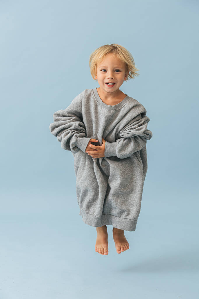 Cute blond toddler boy in a huge oversized grey longsleeve. He's jumping in the air, bending his knees. Over blue background. - Photo, Image