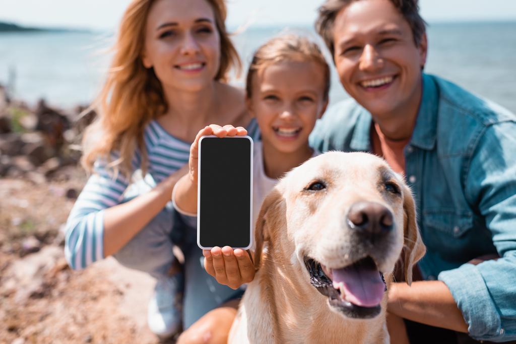 Enfoque selectivo de la mujer sosteniendo teléfono inteligente cerca de la familia y golden retriever en la playa - Foto, Imagen