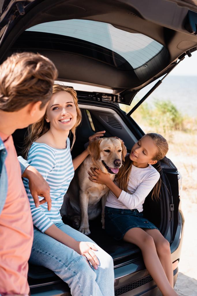 Selective focus of woman looking at husband near daughter and golden retriever in car trunk  - Photo, Image
