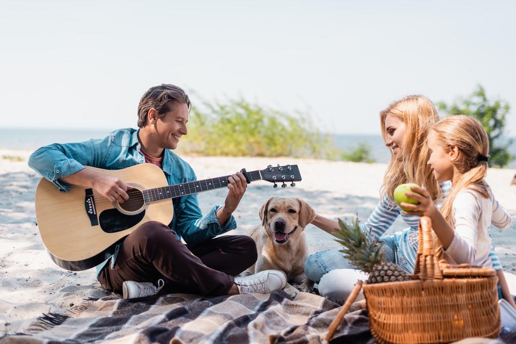家族の近くでアコースティックギターを演奏し、ビーチで黄金のレトリバーの選択的な焦点  - 写真・画像