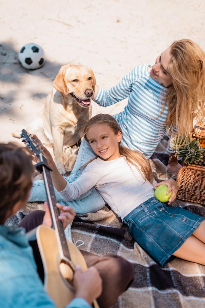 Felülnézet férfi játszik akusztikus gitár közelében feleség, lánya és golden retriever a strandon  - Fotó, kép