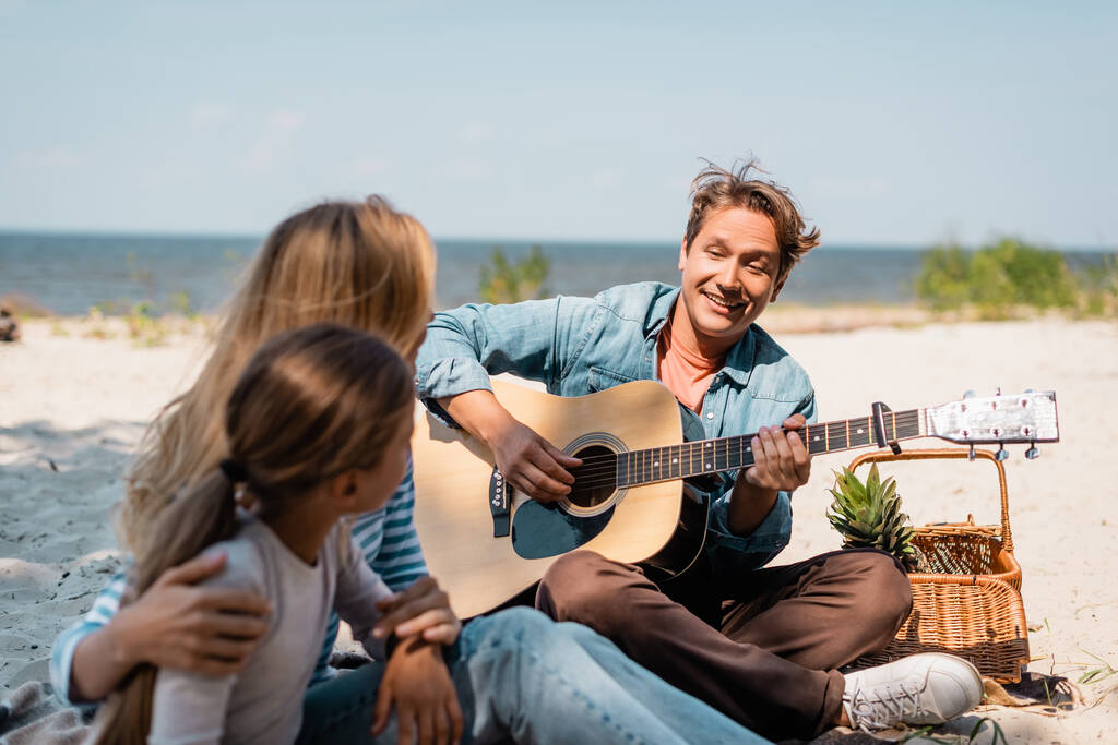 Enfoque selectivo del hombre tocando la guitarra acústica cerca de la esposa y el niño en la playa  - Foto, imagen