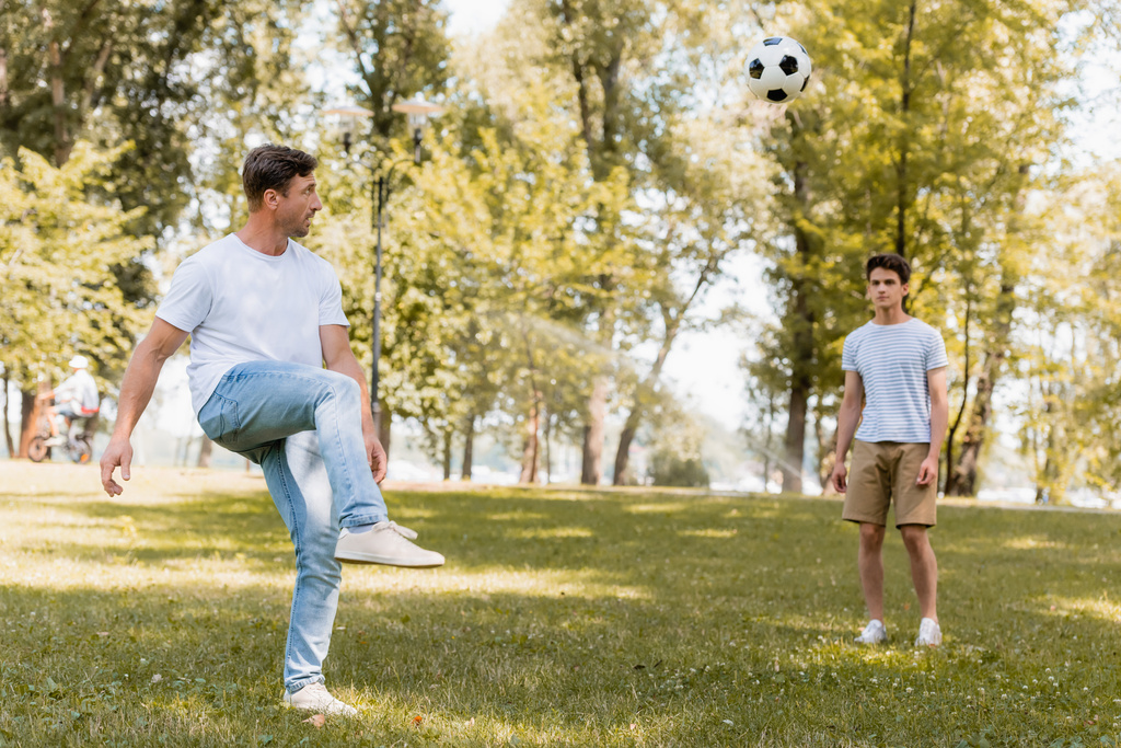 селективное внимание мужчины, играющего в футбол с сыном-подростком в парке - Фото, изображение