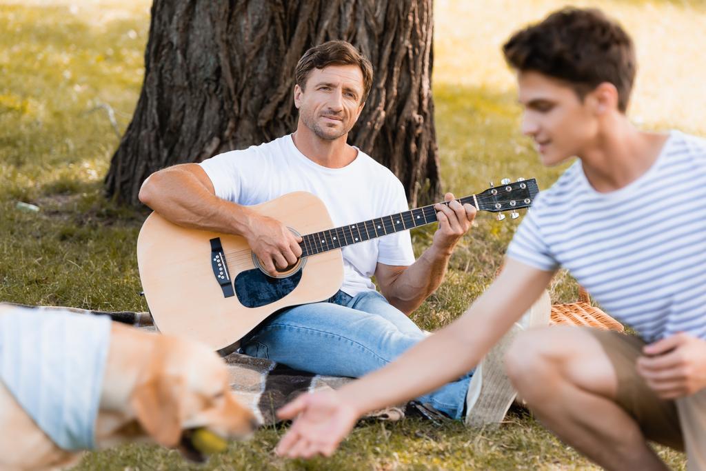 messa a fuoco selettiva dell'uomo seduto sotto l'albero e suonare la chitarra acustica mentre guarda figlio adolescente e golden retriever - Foto, immagini