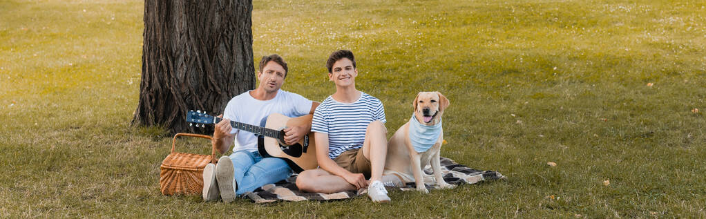 panoramic crop of teenager boy and father sitting on blanket near golden retriever under tree trunk  - Photo, Image