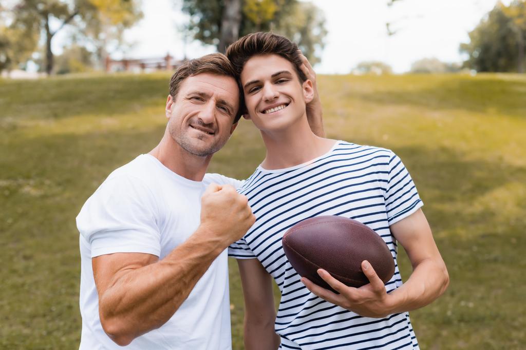 гордый отец, показывающий сжатый кулак и стоящий рядом с сыном-подростком с мячом для регби  - Фото, изображение