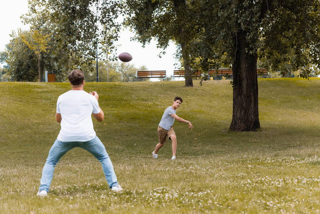 επιλεκτική εστίαση του έφηβος αγόρι παίζει αμερικανικό ποδόσφαιρο με τον πατέρα στο πράσινο πάρκο  - Φωτογραφία, εικόνα