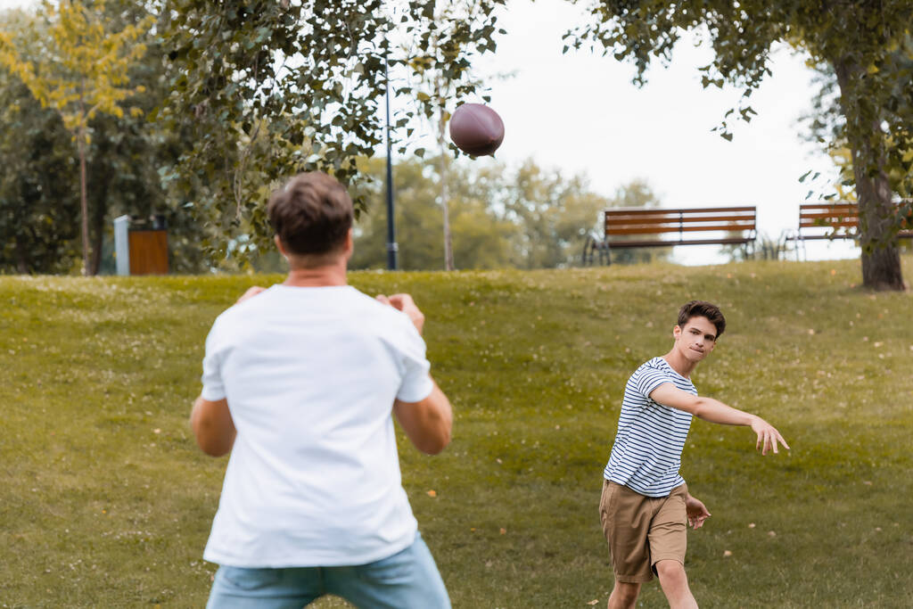selective focus of teenager boy throwing rugby ball to father in park  - Photo, Image