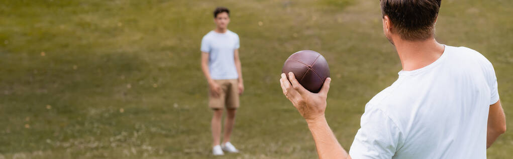 緑の公園で10代の息子の近くでラグビーボールを持っている父親のパノラマの概念  - 写真・画像