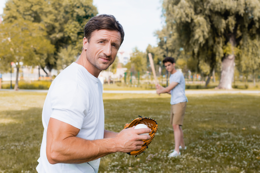 selektywne skupienie człowieka w skórzanej rękawicy patrząc w kamerę i trzymając piłkę grając w baseball z nastoletnim synem w parku  - Zdjęcie, obraz