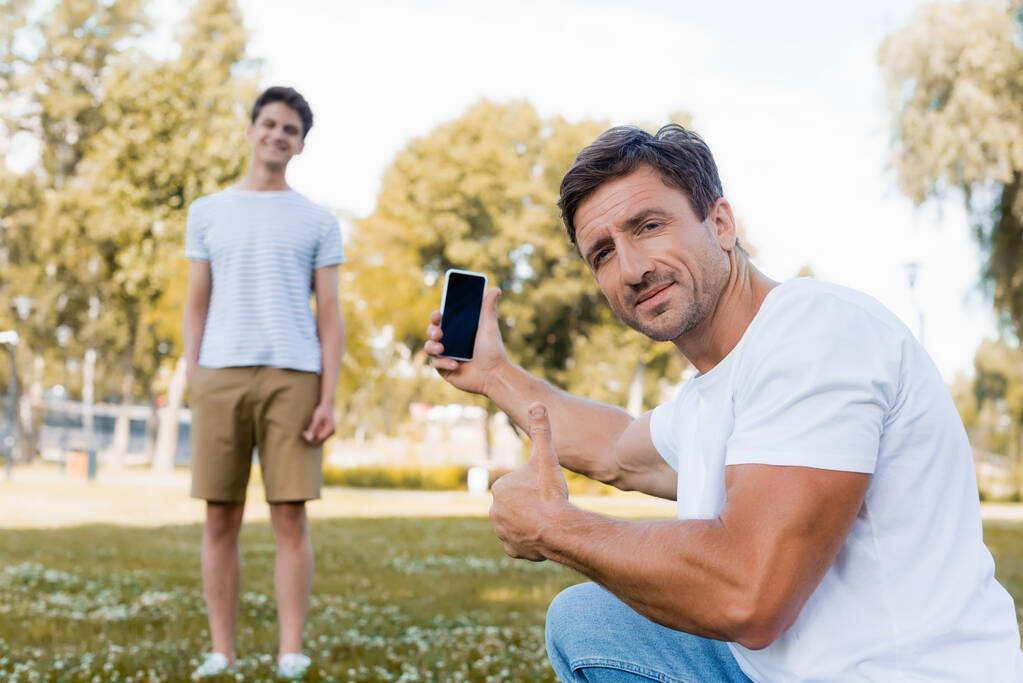избирательный фокус отца, держащего смартфон и показывающего большой палец рядом с сыном-подростком в парке  - Фото, изображение