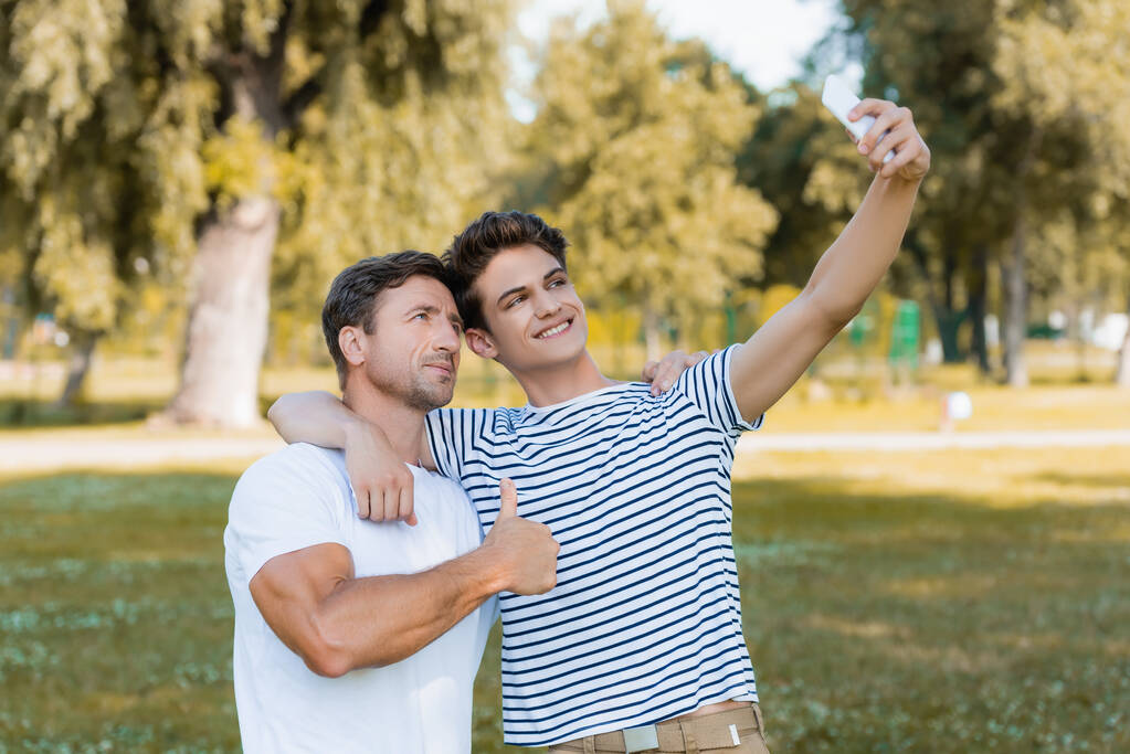 otec ukazující palec nahoru, zatímco teenager syn přičemž selfie v parku  - Fotografie, Obrázek