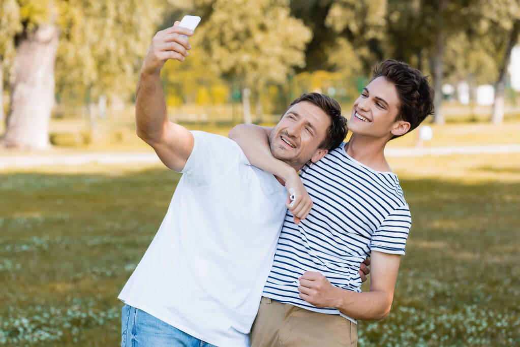 adolescente hijo abrazando padre mientras toma selfie en parque  - Foto, imagen