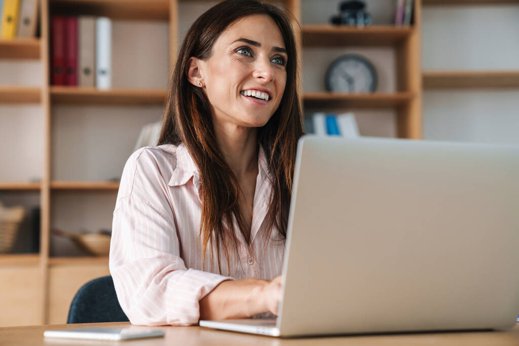 Зображення щасливої красивої бізнес-леді посміхається під час роботи з ноутбуком в офісі
 - Фото, зображення