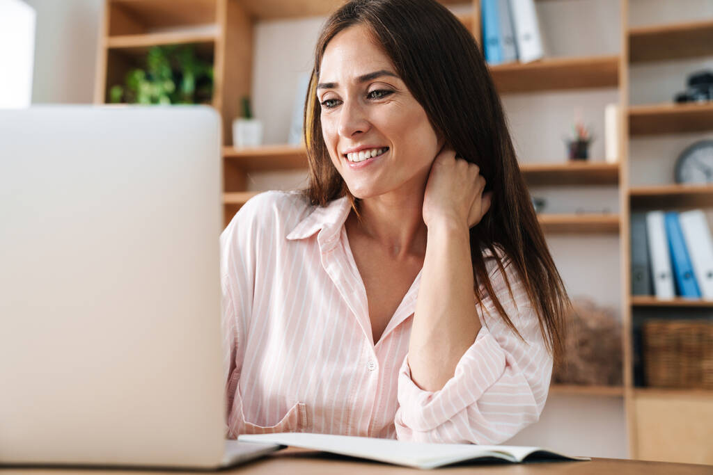 Зображення щасливої дорослої бізнес-леді, яка посміхається під час роботи з ноутбуком в офісі
 - Фото, зображення