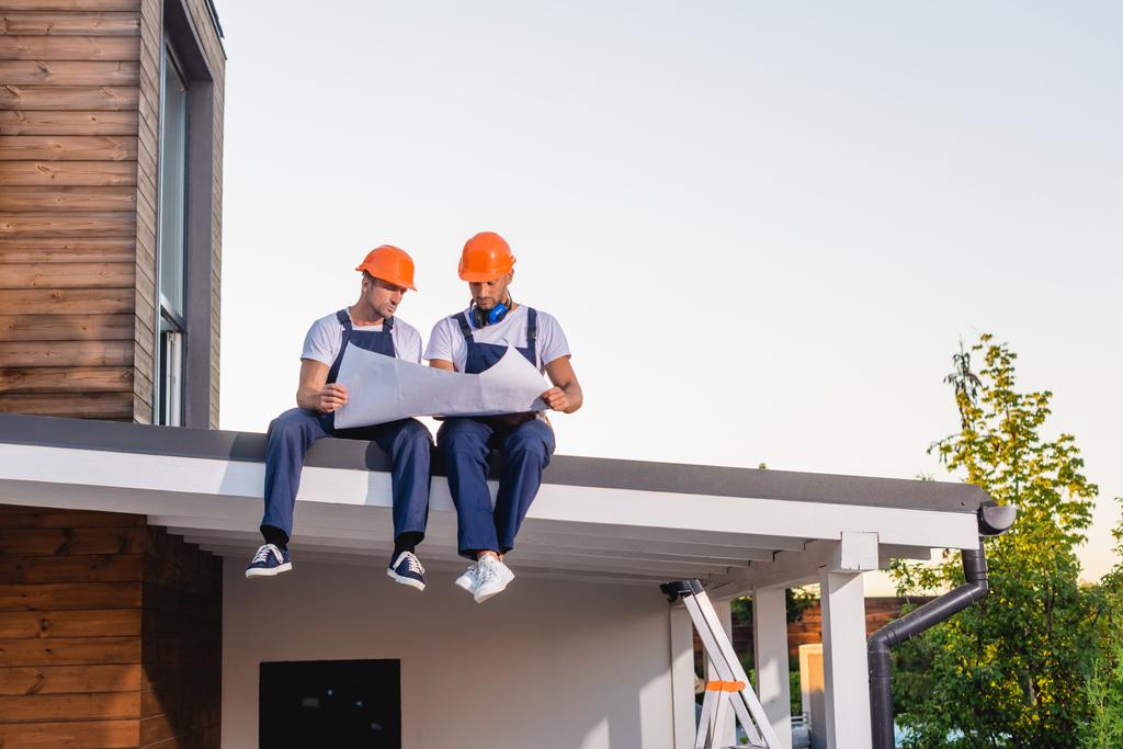 Werknemers in uniform en hardhoeden werken met blauwdruk op het dak van het gebouw  - Foto, afbeelding