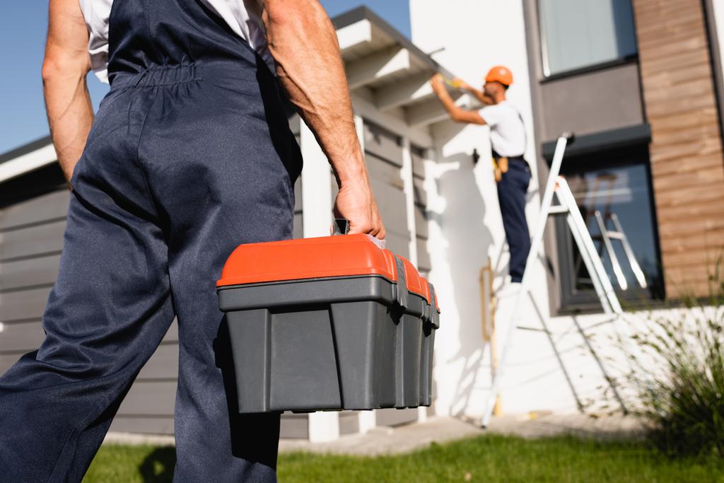 Enfoque selectivo de constructor sosteniendo caja de herramientas mientras colega trabajando cerca de casa en segundo plano  - Foto, Imagen
