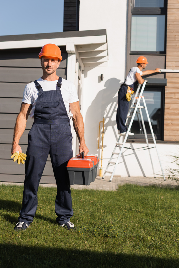 Selektiver Fokus des Bauarbeiters mit Handschuhen und Werkzeugkiste, der auf dem Rasen steht, während der Kollege an der Leiter in der Nähe des Hauses arbeitet  - Foto, Bild