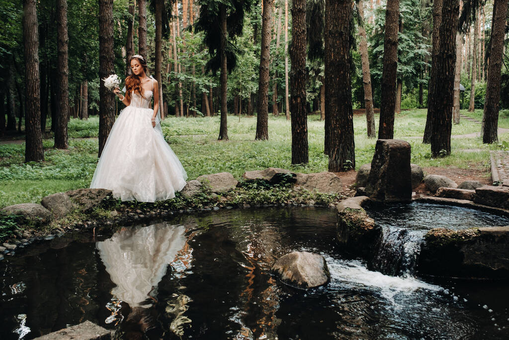 Une mariée élégante dans une robe blanche et des gants tenant un bouquet se tient près d'un ruisseau dans la forêt, profitant de la nature.Un modèle dans une robe de mariée et des gants dans un parc naturel.Biélorussie. - Photo, image