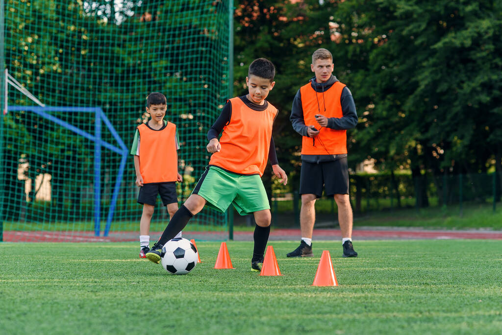 Постійний хлопчик-підліток у спортивному одязі тренує футбол на футбольному полі і вчиться крутити м'яч між тренувальними конусами
. - Фото, зображення