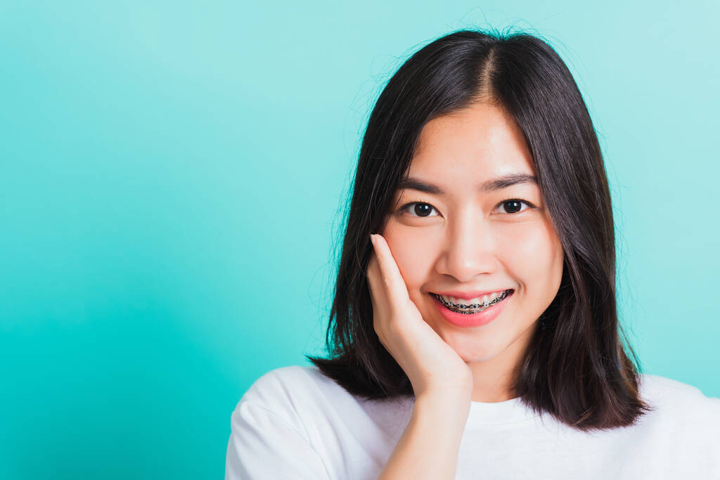 Retrato de adolescente asiática hermosa sonrisa de mujer tiene frenos dentales en los dientes riendo ella tocando su cara a mano, estudio de tiro aislado sobre un fondo azul, Medicina y odontología concepto - Foto, imagen