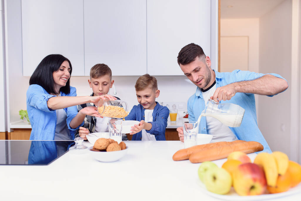 Mama, tata i dwóch synów jedzących zdrowe śniadanie z płatkami kukurydzianymi i świeżym mlekiem na przytulnej kuchni przy stole. - Zdjęcie, obraz