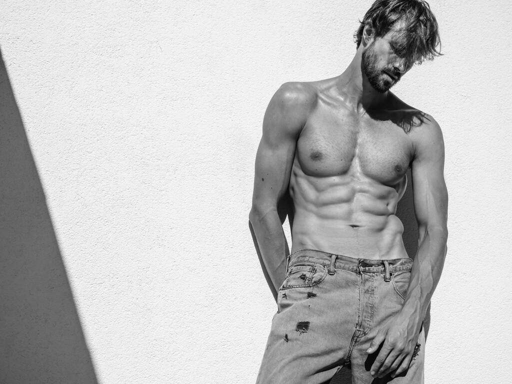 Retrato de un deportista guapo y fuerte. Modelo de fitness deportivo saludable posando cerca de la pared blanca en jeans. Hombre de moda sexy seguro con torso desnudo desnudo. Lambersexual al aire libre - Foto, imagen
