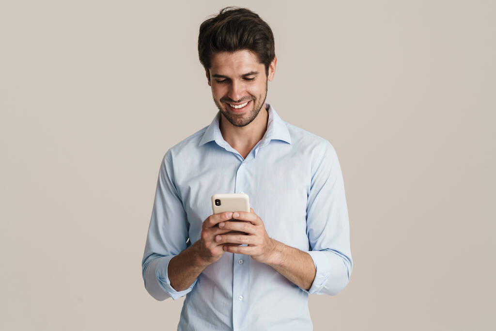 Bild eines glücklichen geschäftstüchtigen brünetten Mannes, der sein Handy in der Hand hält, während er isoliert vor beigem Hintergrund steht - Foto, Bild