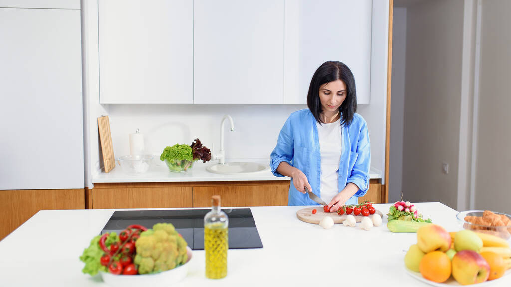 Hermosa mujer corta tomates en una tabla de madera para preparar ensalada saludable de verduras frescas en la cocina acogedora. - Foto, imagen