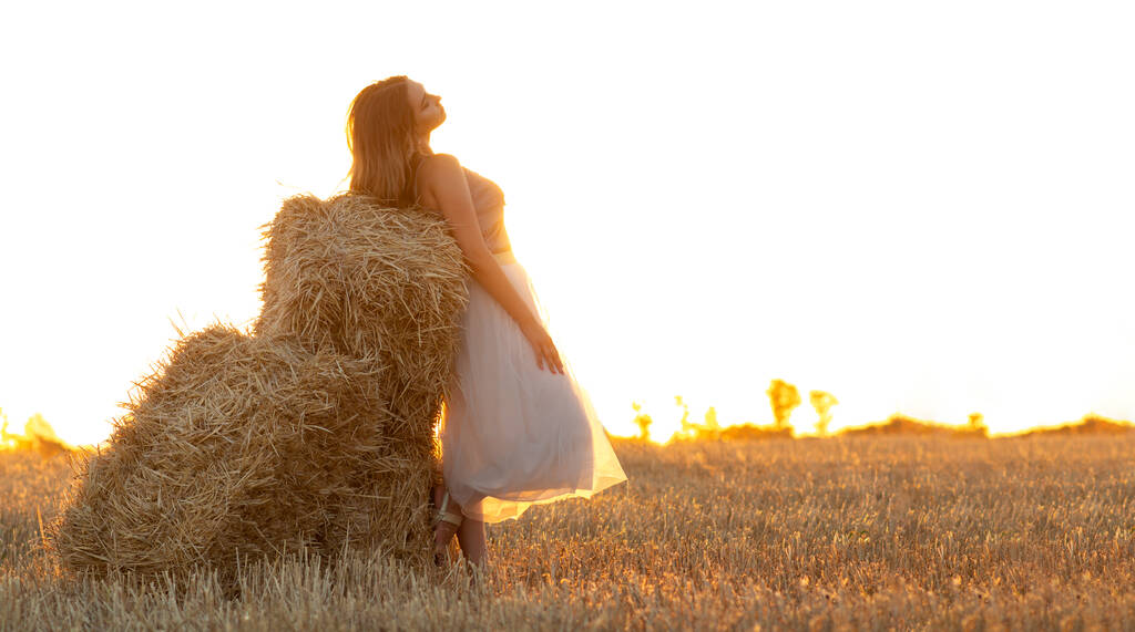Молодая женщина опирается на стог сена ходьба в летний вечер, красивая романтическая девушка с длинными волосами на открытом воздухе на закате - Фото, изображение