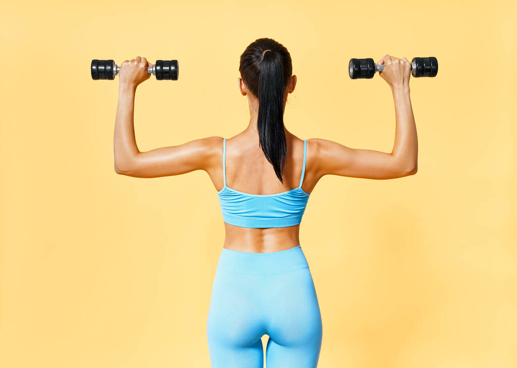 Задний вид спортивной женщины в спортивной одежде, делающей упражнения с гантелями на желтом фоне - Фото, изображение