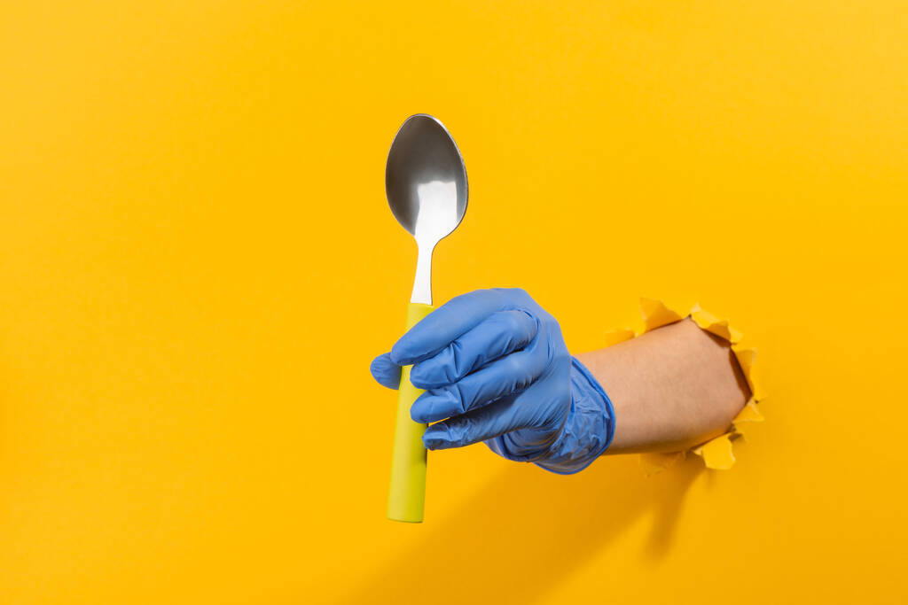 Main dans un gant en latex donnant une cuillère en métal à travers le mur jaune déchiré - Photo, image