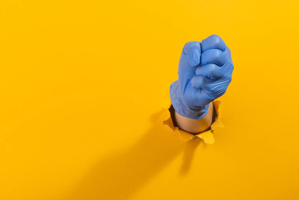 Pięść w lateksowej rękawiczce przebija się przez rozdartą żółtą ścianę - Zdjęcie, obraz