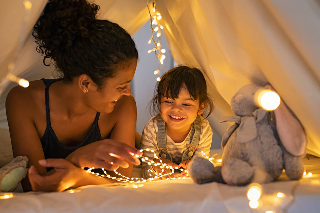 Playful mother and her daughter playing in children room in lighted tent. African mom and lovely cute girl having fun in kid hut at night at home. Mother and little daughter spending time together with strip lights. - Photo, Image