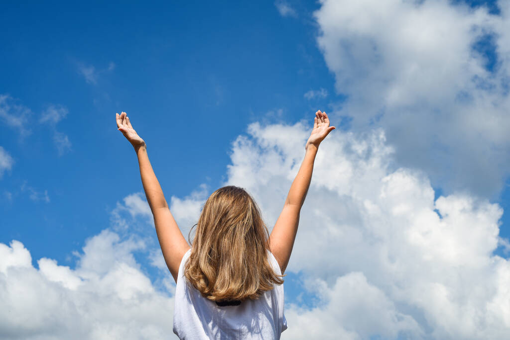 een vrouw of meisje staat met haar rug naar de camera en strekt haar handen naar de hemel. Het meisje hief haar handen op tegen de blauwe lucht. - Foto, afbeelding