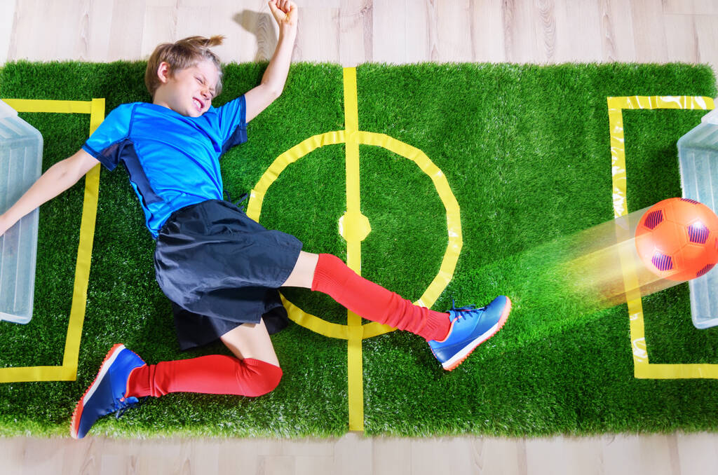 Мальчик имитирует футбольный матч с ударом мячом по зеленому полю возле экрана телевизора во время карантина. Сиди дома. Спортивные мероприятия. Ситуация с коронавирусом. Вид сверху - Фото, изображение